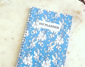 May include: A blue spiral bound notebook with a white floral pattern and the text "IVF Planner" on the cover.