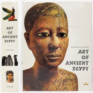May include: A book cover with a close-up of a wooden sculpture of a person's face. The book title is "Art of Ancient Egypt" by Kazimierz Michalowski. The book is published by Abrams.