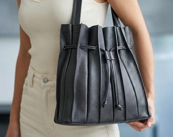 May include: Black leather tote bag with a pleated design and drawstring closure. The bag has a shoulder strap and is held by a person. The bag is a dark color and appears to be made of textured leather.