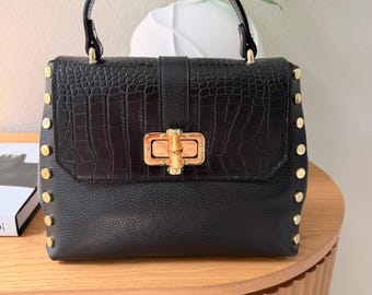 May include: Black leather handbag with a top handle and gold-colored hardware. The flap features a crocodile-embossed design. Gold studs line the sides of the bag. The bag is sitting on a wooden table.