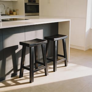 May include: Two black wooden bar stools with a curved seat and a four-legged base. The stools are positioned in front of a kitchen island with a light-colored countertop. The stools have a rustic, distressed finish.