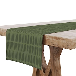 May include: Olive green table runner with a repeating teardrop pattern in a darker shade of green. The runner is draped over a wooden table, showcasing its design and texture. The table has a rustic, natural wood finish.