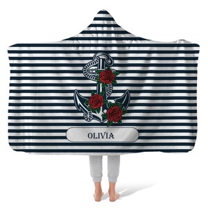 May include: A hooded blanket with a navy blue and white striped pattern. A large anchor with red roses is centered on the blanket. The name "OLIVIA" is printed in a white rectangle below the anchor.