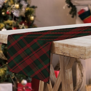 May include: A red and green plaid table runner is draped over a wooden table. The runner features a classic tartan pattern with intersecting lines of red and dark green. The table runner is a festive decoration for the holiday season.