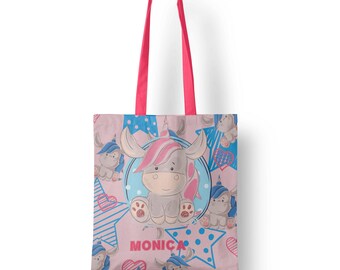May include: Pink tote bag with a repeating pattern of cartoon unicorns, stars, and hearts in blue and pink. The bag has pink handles and the name "MONICA" is printed on the front.