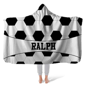 May include: A hooded blanket featuring a black and white soccer ball pattern with a central horizontal white band displaying the name RALPH in bold black capital letters. The blanket has a pointed hood and a rectangular shape with rounded corners.
