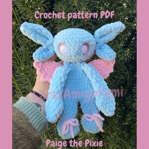 Könnte beinhalten: Eine handgefertigte blaue Plüsch-Pixie-Puppe mit rosa Flügeln und Akzenten. Die Puppe hat große Augen mit Glitzer und rosa Schleifen an den Füßen. Der Text "Crochet pattern PDF" und "Paige the Pixie" sind sichtbar.