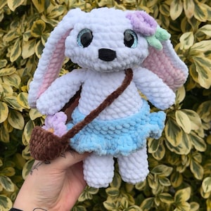 Könnte beinhalten: Ein weißer Häkelhase mit blauen Augen, einem hellblauen Rock und einer braunen Häkeltasche mit lila Blumen. Der Hase hält einen braunen Riemen.