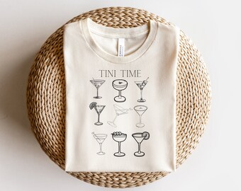 Könnte beinhalten: Ein beiges T-Shirt mit dem Aufdruck "TINI TIME" auf der Vorderseite. Das Shirt zeigt ein Design aus neun verschiedenen Martini-Gläsern.