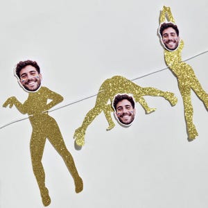 May include: A decorative banner featuring three gold glitter cutouts of human figures in various poses, each topped with a smiling face cutout of the same person with rainbow face paint, all strung together on a thin white string.