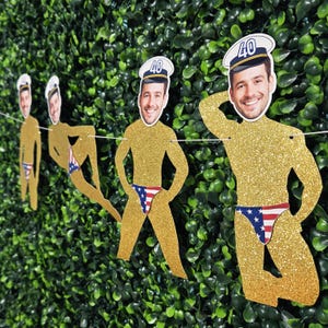 May include: A gold glitter banner featuring cutouts of a man in a sailor hat and American flag speedo. Each cutout has a face and the number "40" on the hat. The banner is strung on a white string and is displayed against a green hedge.
