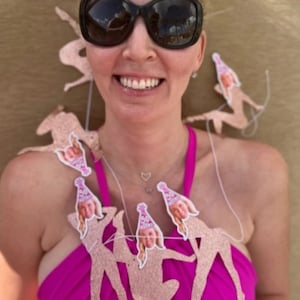 woman celebrates her birthday with her face banner adorned around her neck, she smiles at the camera in a bathing suit while on the beach