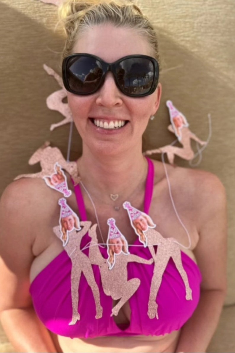 woman celebrates her birthday with her face banner adorned around her neck, she smiles at the camera in a bathing suit while on the beach