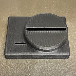 May include: A black 3D-printed object with a rectangular base and a circular top. The top has a long, narrow slot cut through it. The object appears to be made of ridged plastic.