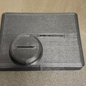 May include: A black plastic rectangular box with two slots. The slots are rectangular and have a thin, horizontal line cut into them.
