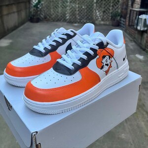 May include: White, orange, and black custom Nike Air Force 1 sneakers with a cartoon cowboy graphic on the side.