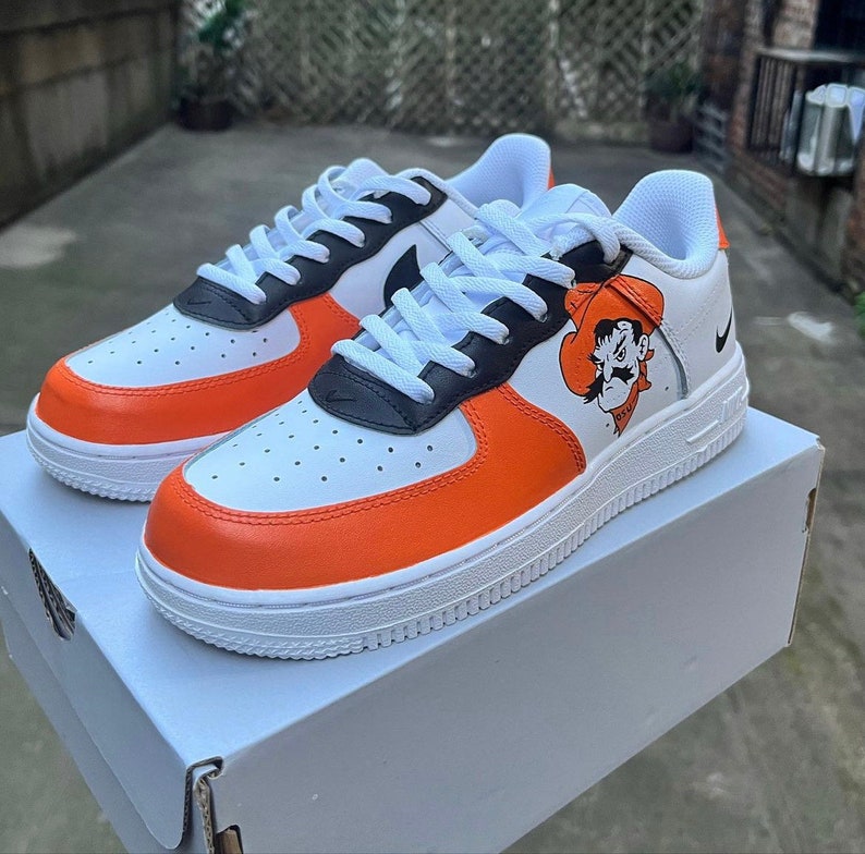 May include: White, orange, and black custom Nike Air Force 1 sneakers with a cartoon cowboy graphic on the side.