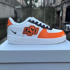 May include: White leather sneakers with an orange and black OSU logo. The shoes have a white midsole and a black outsole.