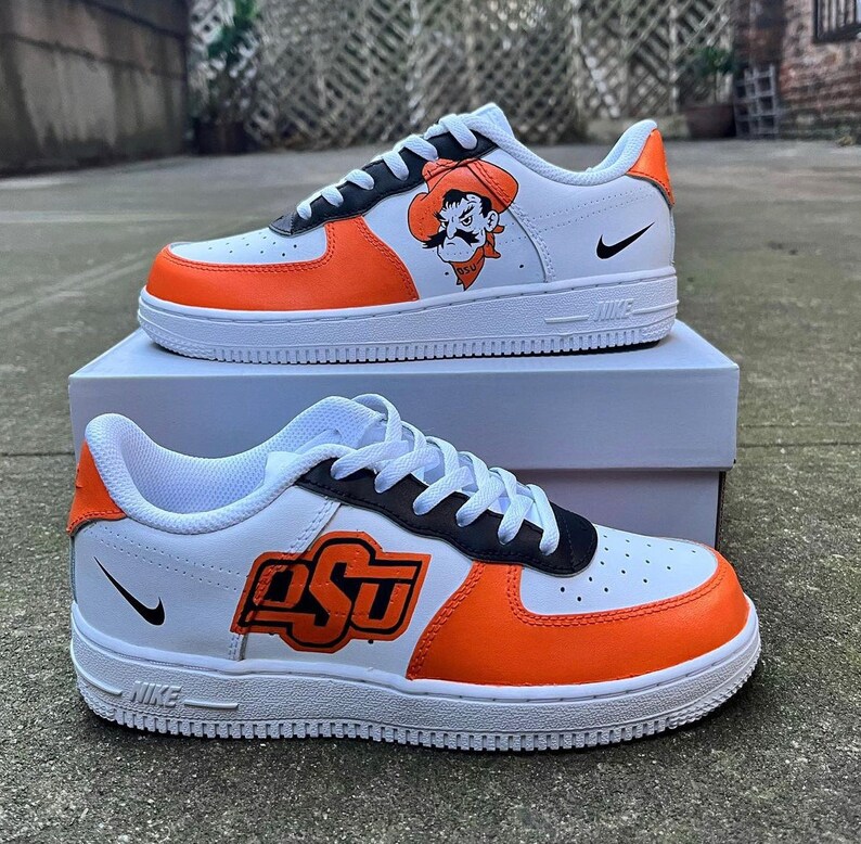 May include: A pair of white Nike Air Force 1 sneakers with orange and black accents. The sneakers feature a custom design with the letters "OSU" and a cartoon mascot wearing a cowboy hat.