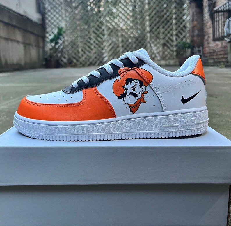 May include: White, orange, and black Nike Air Force 1 sneakers with a custom design featuring an orange and black mascot with a cowboy hat and the text "OSU".