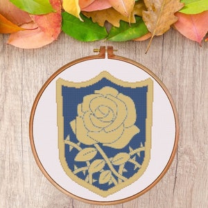 Puede incluir: Un diseño bordado en oro y azul de una rosa con espinas sobre un fondo blanco. El diseño tiene forma de escudo y está enmarcado en un aro de bordado de madera.