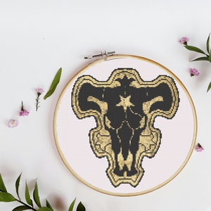 Puede incluir: Diseño de punto de cruz bordado en negro y dorado de una cabeza de toro con una estrella de cinco puntas en el centro, enmarcado en un aro de madera redondo.