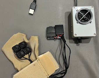 May include: A beige fabric glove with four black buttons, a beige velcro strap, and a black USB cable connected to a small black box with a red wire. The box is connected to a larger gray box with a black speaker and a black knob.