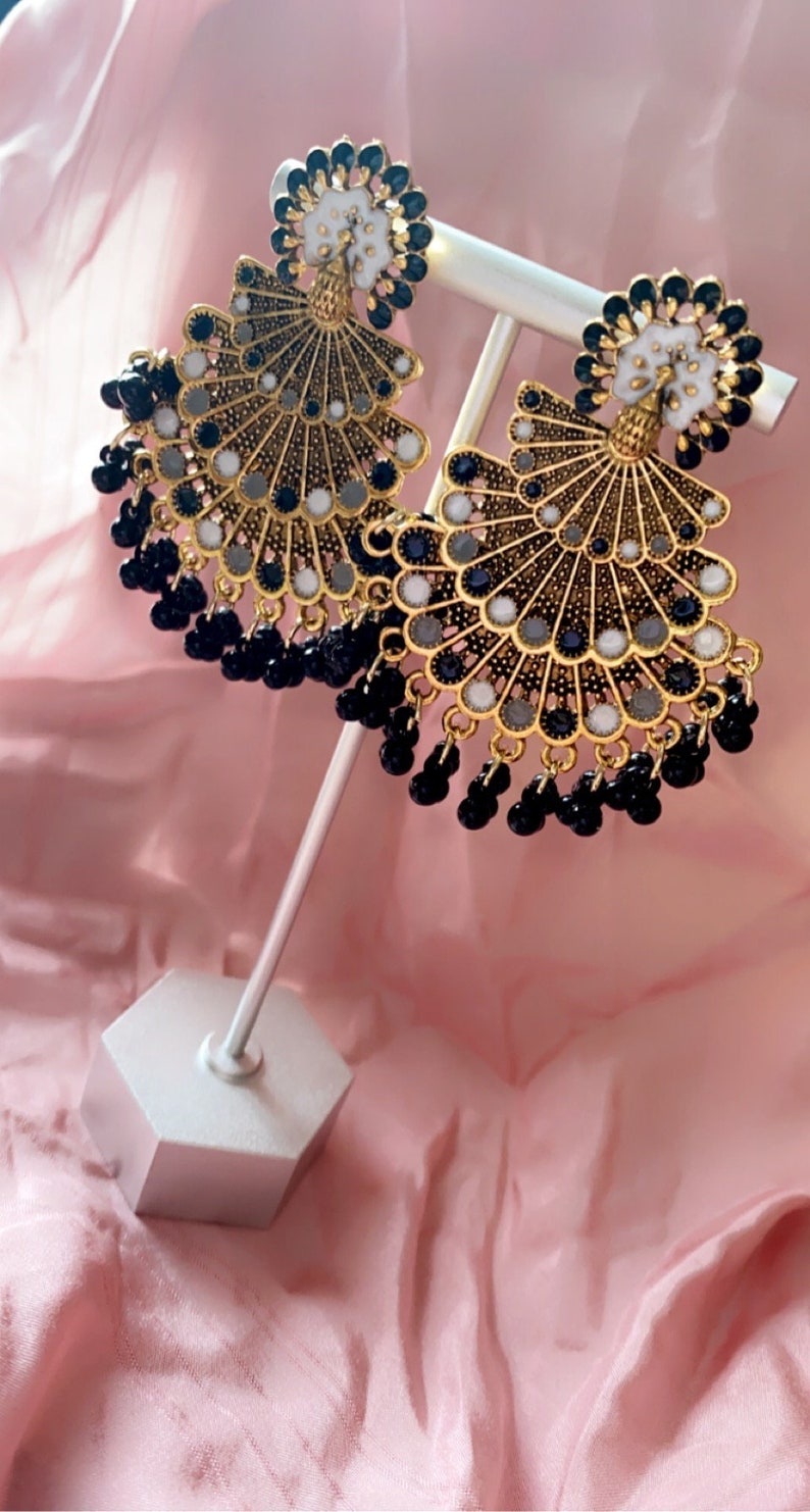 May include: A pair of gold-tone earrings with black and white beads. The earrings feature a peacock design with a fan-shaped bottom and a round top. The earrings are hanging from a white stand.