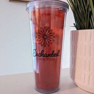 May include: A clear plastic tumbler with a red beverage inside. The tumbler has a black daisy design and the text "Enchanted Loaded Teas" printed on the side.