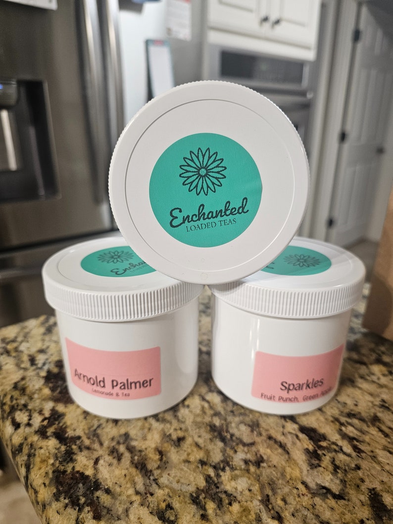 Op de afbeelding: Drie witte containers met deksels, gestapeld op een aanrecht. Elke container heeft een roze label met tekst. De bovenste container heeft een turquoise cirkel met een bloemmotief en de woorden "Enchanted Loaded Teas". De andere containers zeggen "Arnold Palmer" en "Sparkles".
