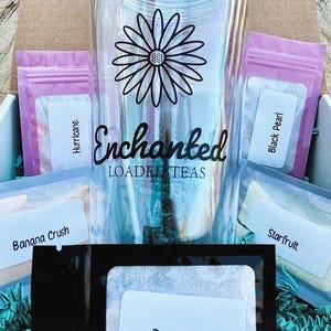 May include: A clear tumbler with a black daisy design and the words "Enchanted Loaded Teas." The tumbler is surrounded by small packets of tea with labels like "Hurricane," "Black Pearl," and "Berry Blast." The items are in a cardboard box with blue crinkle paper.