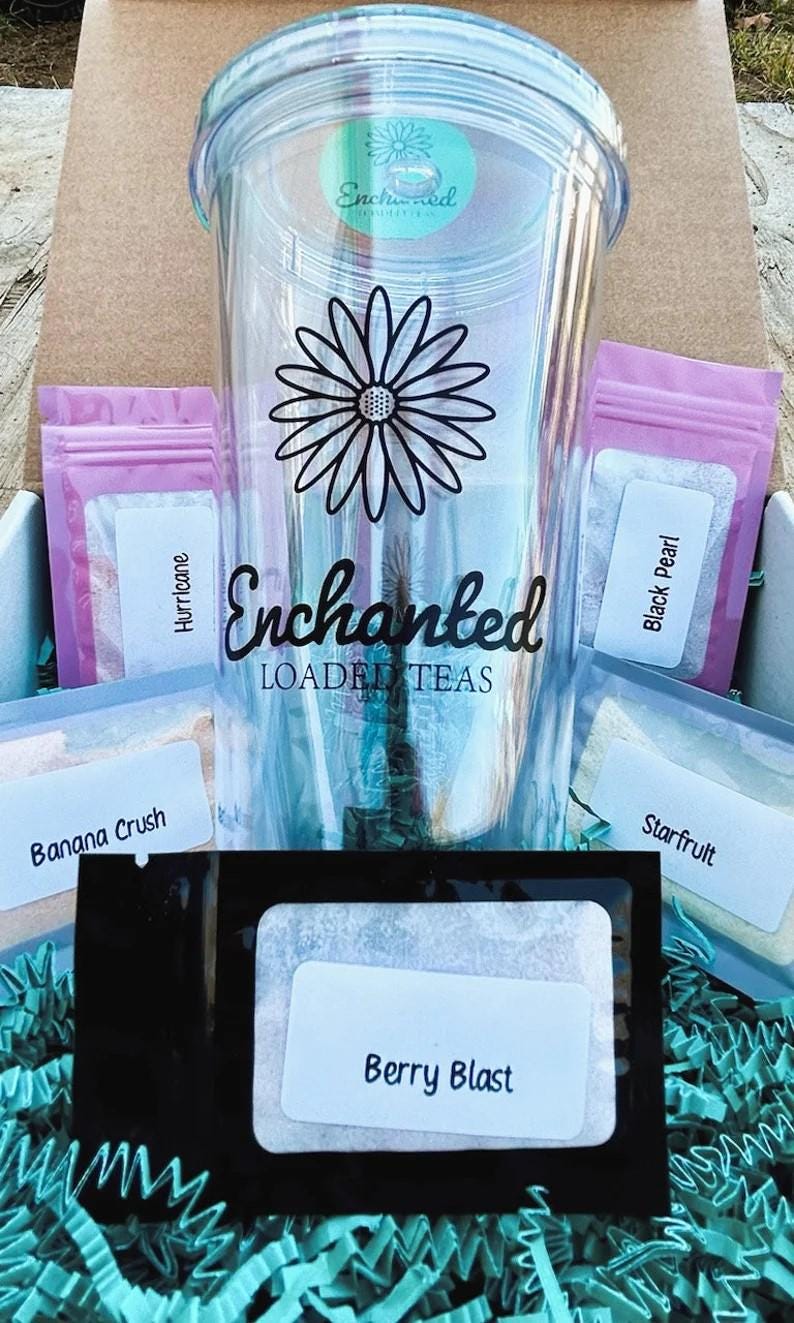 May include: A clear tumbler with a black daisy design and the words "Enchanted Loaded Teas." The tumbler is surrounded by small packets of tea with labels like "Hurricane," "Black Pearl," and "Berry Blast." The items are in a cardboard box with blue crinkle paper.
