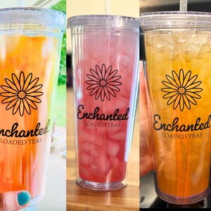 May include: Three clear plastic cups with lids and straws filled with different colored beverages. The cups have a black and white daisy design and the text "Enchanted Loaded Teas".
