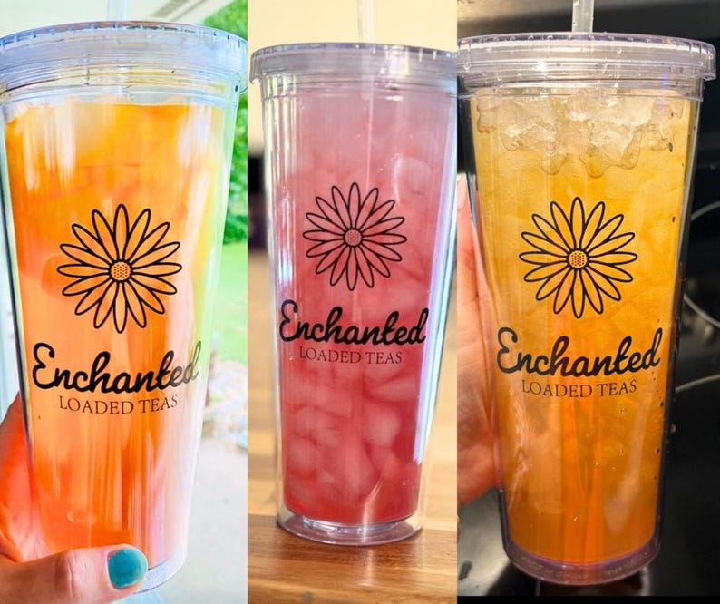 May include: Three clear plastic cups with lids and straws filled with different colored beverages. The cups have a black and white daisy design and the text "Enchanted Loaded Teas".