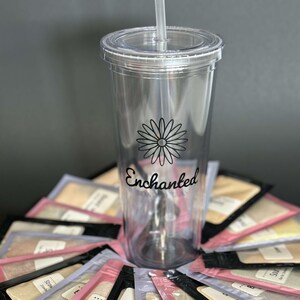 Puede incluir: Un vaso transparente de pl&aacute;stico con una margarita negra y la palabra "Enchanted" impresa en el lateral. El vaso est&aacute; rodeado de peque&ntilde;os paquetes de polvo de colores.