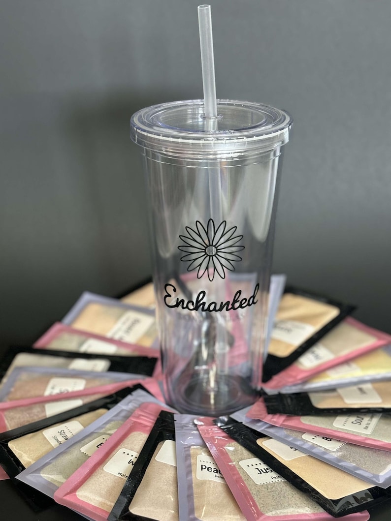Puede incluir: Un vaso transparente de pl&aacute;stico con una margarita negra y la palabra "Enchanted" impresa en el lateral. El vaso est&aacute; rodeado de peque&ntilde;os paquetes de polvo de colores.