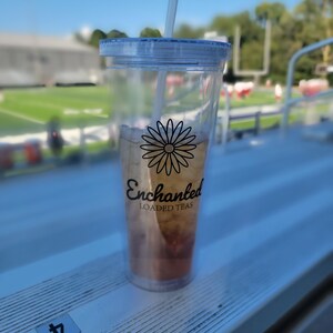 May include: A clear plastic tumbler with a lid and straw, filled with a light brown beverage. The tumbler has a black daisy graphic and the words "Enchanted Loaded Teas" printed on it. The tumbler is sitting on a white surface.