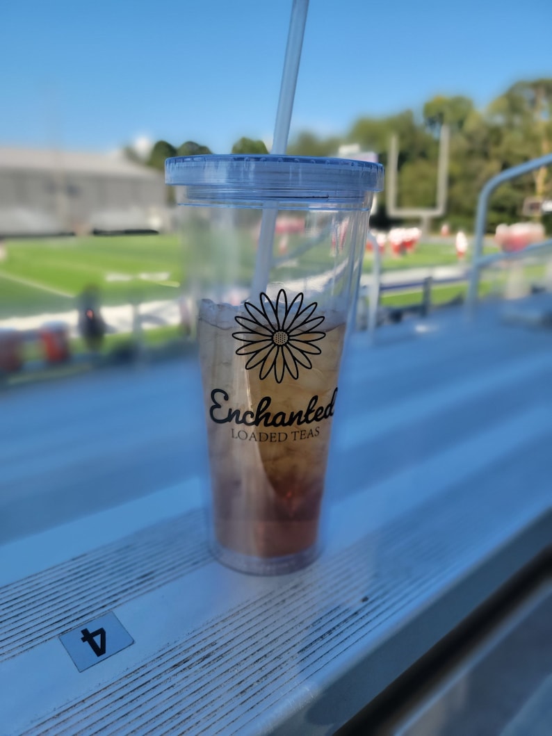 May include: A clear plastic tumbler with a lid and straw, filled with a light brown beverage. The tumbler has a black daisy graphic and the words "Enchanted Loaded Teas" printed on it. The tumbler is sitting on a white surface.