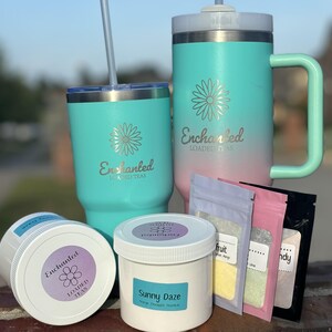 May include: A collection of teal and pink insulated tumblers and jars with the text "Enchanted Loaded Teas". The image also includes small packets of tea and a jar labeled "Sunny Daze".