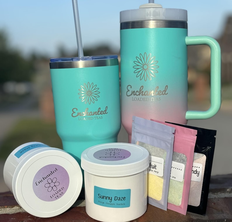 May include: A collection of teal and pink insulated tumblers and jars with the text "Enchanted Loaded Teas". The image also includes small packets of tea and a jar labeled "Sunny Daze".
