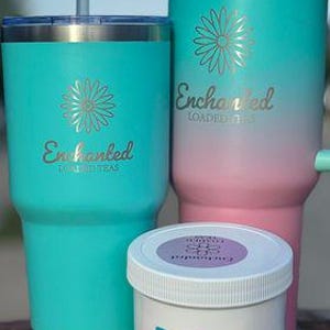 May include: Three insulated tumblers and a small jar. The tumblers are teal and pink with a gold flower design and the word "Enchanted" in gold script. The jar is white with a blue label that says "Sunny Daze".