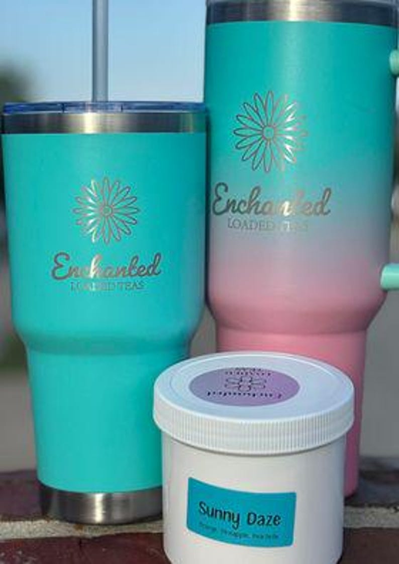 May include: Three insulated tumblers and a small jar. The tumblers are teal and pink with a gold flower design and the word "Enchanted" in gold script. The jar is white with a blue label that says "Sunny Daze".
