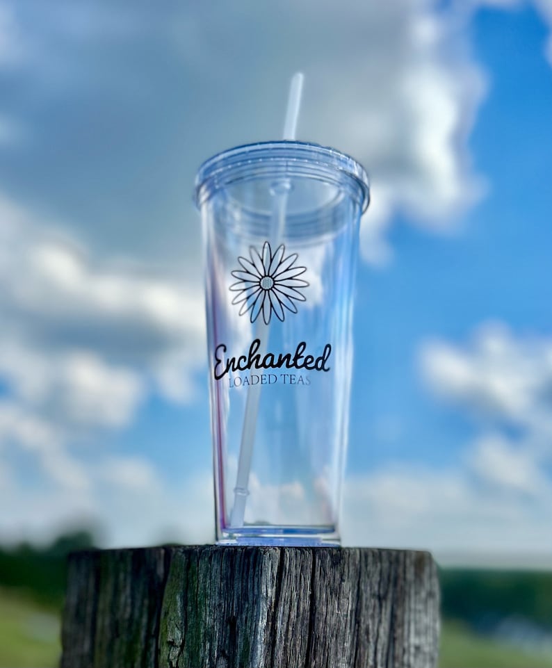 May include: A clear plastic tumbler with a straw, featuring a black and white daisy design and the text "Enchanted Loaded Teas".