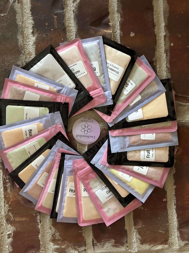 May include: A circular arrangement of small, colorful tea packets. Each packet is sealed and labeled with flavors like "Strawberry," "Peach," and "Watermelon." The packets are in pink, black, and clear packaging, arranged on a brick surface.