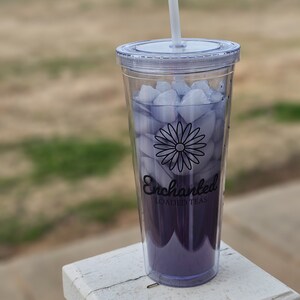 Op de afbeelding: Een doorzichtige plastic beker gevuld met paarse vloeistof en ijsblokjes, met een wit rietje. De beker heeft een zwarte madeliefjesafbeelding en de tekst "Enchanted LOADED TEAS". De drank staat op een wit oppervlak.