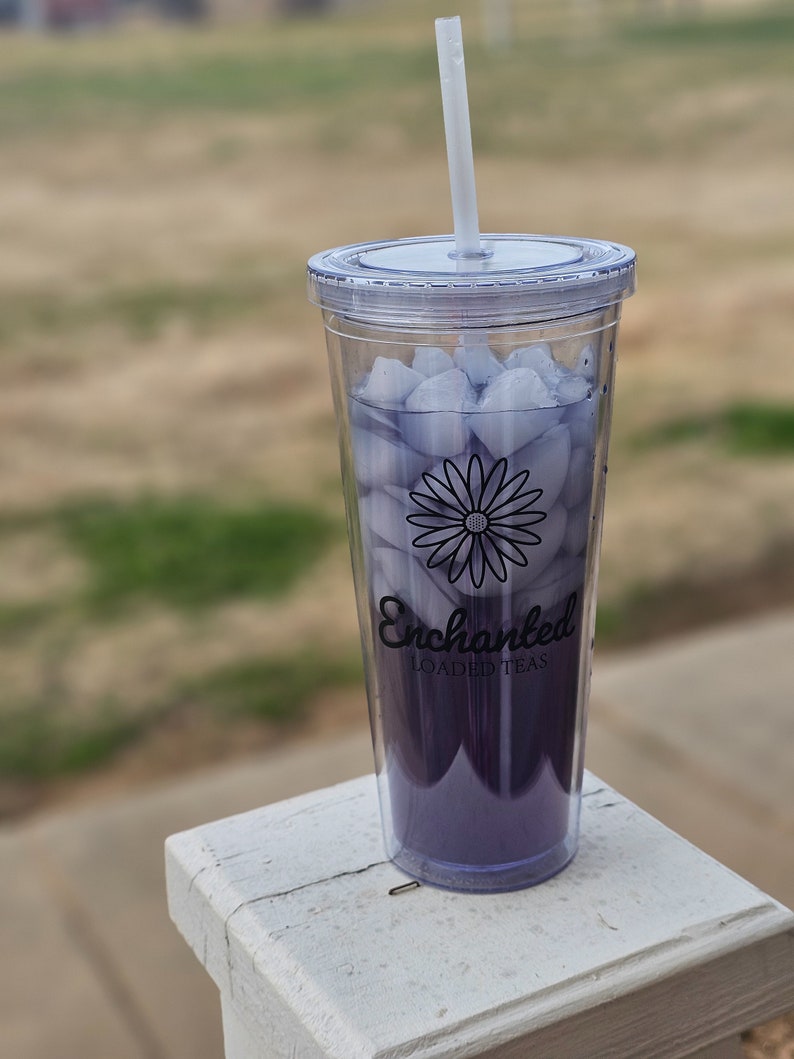Op de afbeelding: Een doorzichtige plastic beker gevuld met paarse vloeistof en ijsblokjes, met een wit rietje. De beker heeft een zwarte madeliefjesafbeelding en de tekst "Enchanted LOADED TEAS". De drank staat op een wit oppervlak.