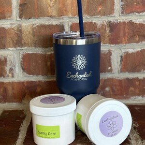 Puede incluir: Un vaso azul de acero inoxidable con una margarita blanca y el texto "Enchanted Loaded Teas" en el lateral. Dos recipientes blancos con etiquetas que dicen "Enchanted Loaded Teas" y "Sunny Daze" est&aacute;n delante del vaso.