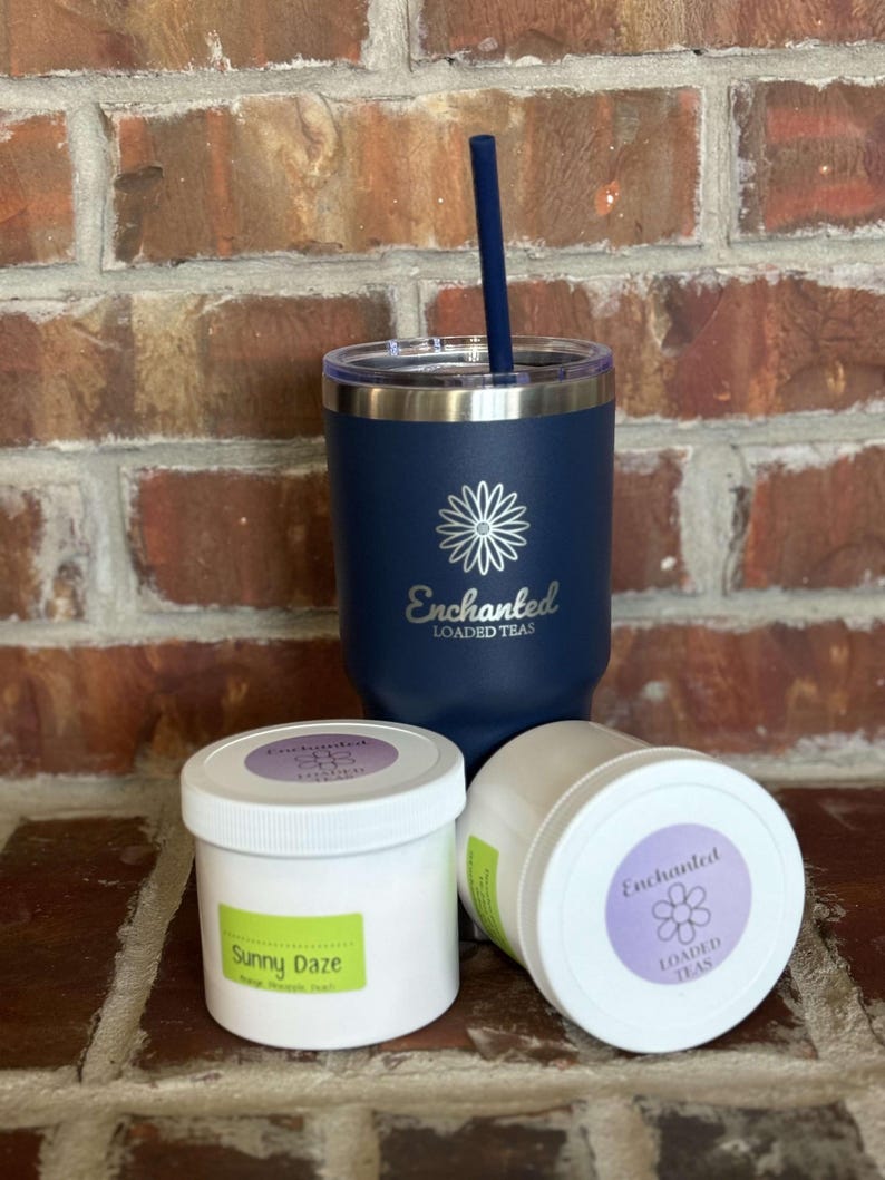 Puede incluir: Un vaso azul de acero inoxidable con una margarita blanca y el texto "Enchanted Loaded Teas" en el lateral. Dos recipientes blancos con etiquetas que dicen "Enchanted Loaded Teas" y "Sunny Daze" est&aacute;n delante del vaso.