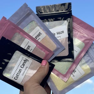 Puede incluir: Cinco peque&ntilde;as bolsas resellables llenas de purpurina iridiscente. Las bolsas est&aacute;n etiquetadas con los nombres "Cotton Candy", "Pops", "Sunset", "Purple Haze" y "Drift".