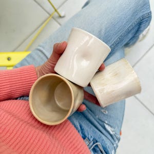 Op de afbeelding: Drie keramische mokken in verschillende tinten wit en beige. De mokken zijn handgemaakt en hebben een unieke, textuur.